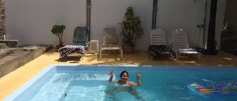 Una piscina con una persona haciendo el gesto de la paz, rodeada de sillones de descanso y plantas en macetas.