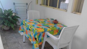 Un pequeño comedor con mantel colorido, sillas de plástico blanco, una planta en maceta y una ventana con vistas al exterior.