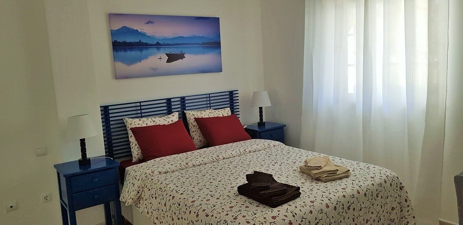 Un dormitorio con una cama, dos mesitas de noche, una lámpara, un cuadro y una ventana con cortinas.