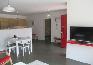 Cocina moderna con armarios blancos, una mesa de comedor con sillas, un televisor sobre un mueble rojo e iluminación colgante.