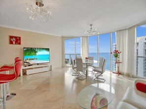 Sala de estar moderna com televisão grande, cadeiras vermelhas, mesa de centro de vidro e vista para o mar.