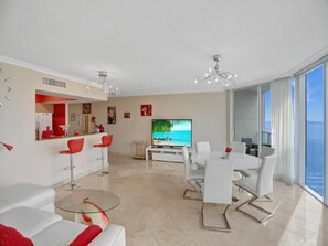 Uma sala de estar moderna com televisão, móveis brancos, uma mesa de centro de vidro e vista para o mar.