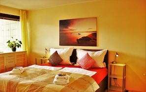 Quarto de hotel com cama, almofadas, mesa de cabeceira, abajur, janela com cortinas e um quadro na parede.