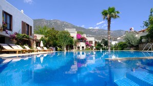 A swimming pool with clear blue water, surrounded by lounge chairs and palm trees, with mountains in the background. - Town Centre Penthouse Apartment With Two Sea View Private Roof Terraces (Kalkan)