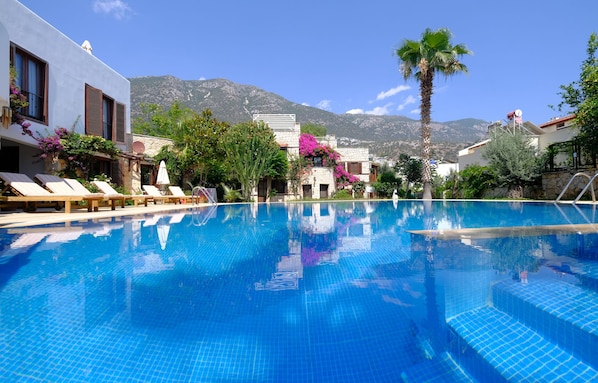 A swimming pool with clear blue water, surrounded by lounge chairs and palm trees, with mountains in the background.