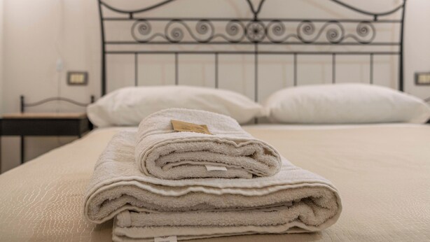 A neatly made bed with a folded white towel on top, a metal headboard, and a bedside table with a lamp.