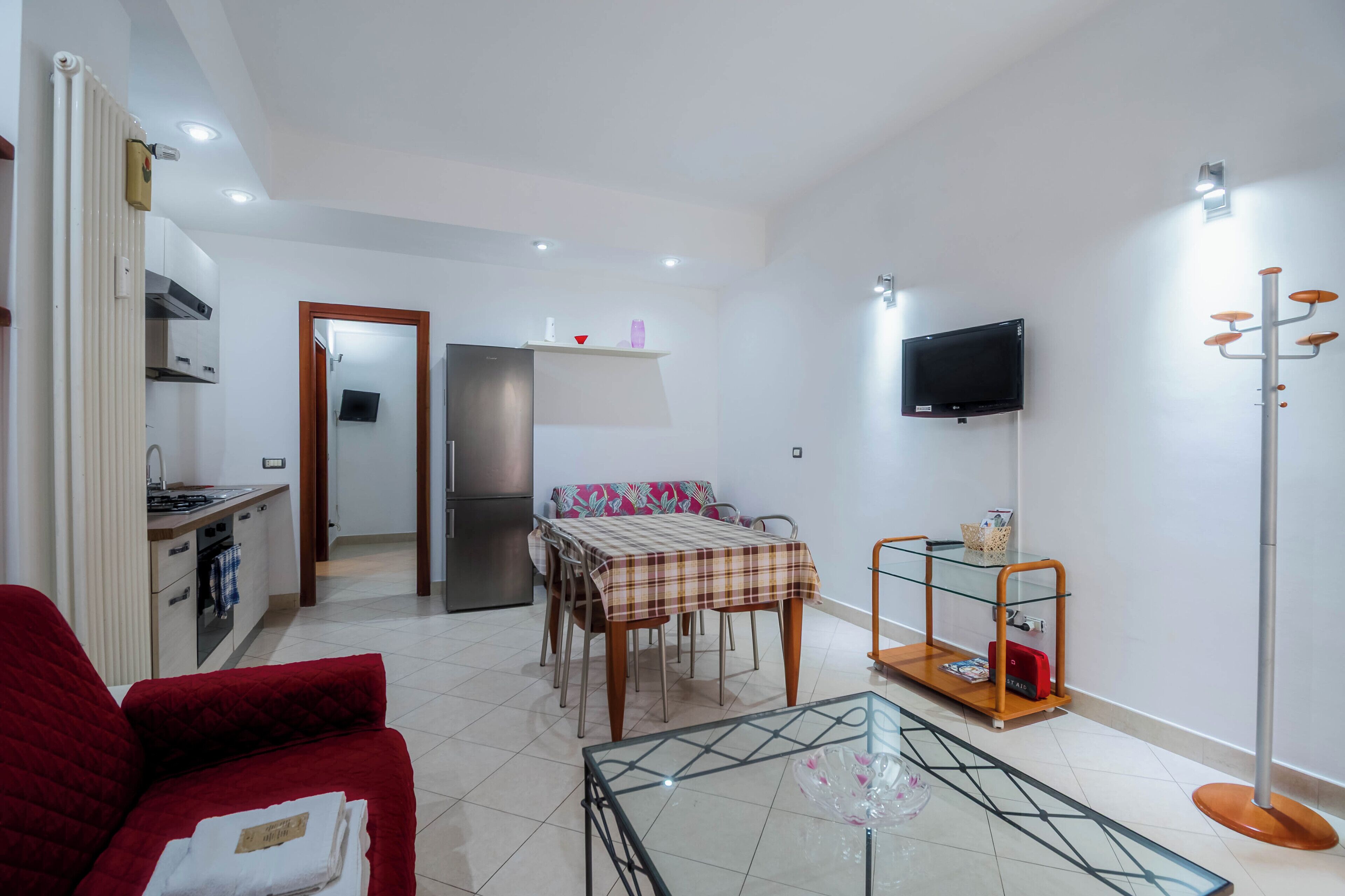 A compact living space with a kitchenette, a dining area with a table and chairs, a red sofa, a glass coffee table, and a TV mounted on the wall.