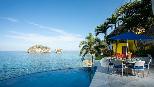 A poolside area with a table and chairs, a thatched-roof structure, and a view of the sea with two small islands. - Great RATES with full STAFF!  Relax and enjoy! (PUERTO VALLARTA)