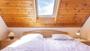 Une chambre avec un plafond en pente, un lambris en bois, un puits de lumière, un lit avec une literie blanche, deux tables de chevet avec des lampes et une petite table de nuit.