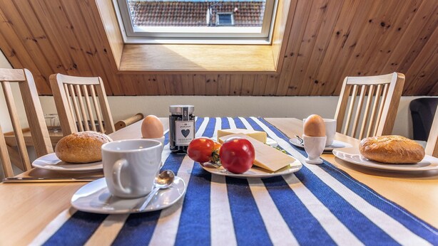 Une table à manger, dressée avec du pain, des œufs, du fromage et des tomates, se trouve dans une pièce aux murs en bois et dotée d’une lucarne.
