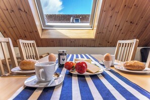 Une table à manger, dressée avec du pain, des œufs, du fromage et des tomates, se trouve dans une pièce aux murs en bois et dotée d’une lucarne.