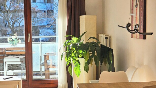A modern dining area with a wooden table and white chairs, a potted plant, and a view through a glass door to the outside. - Stylish bright apartment, very conveniently. Metro just around the corner. Internet (München)