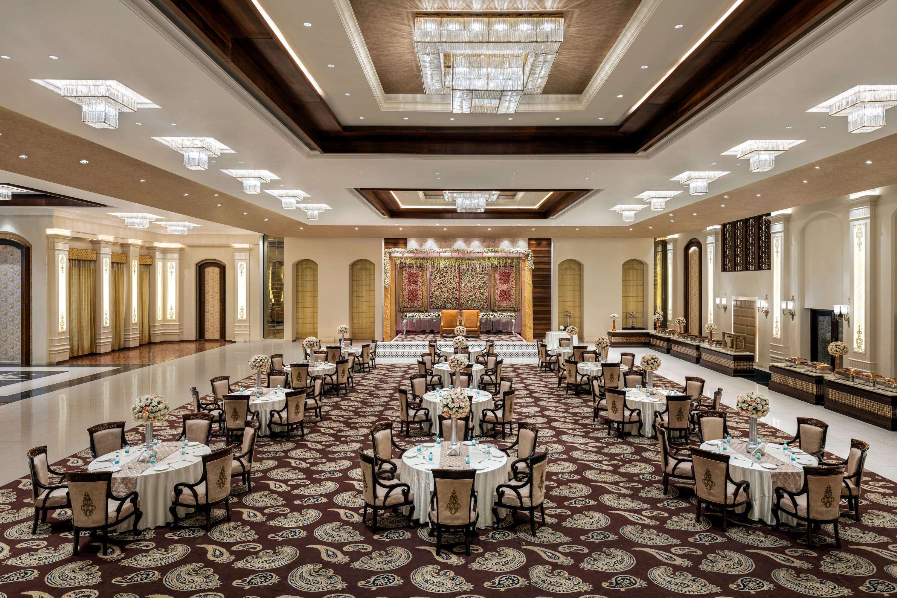 A spacious banquet hall with round tables, chandeliers, and a patterned carpet.