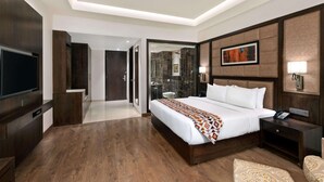 Quarto de hotel moderno com uma cama grande, televisão de tela plana, mesa de cabeceira com telefone e banheiro visível por uma porta de vidro.