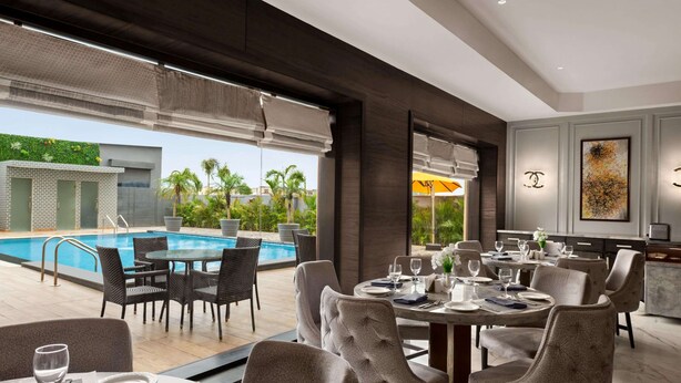 Uma área de jantar moderna com vista para a piscina, mesas de jantar postas com pratos e copos, e uma grande janela com persianas.