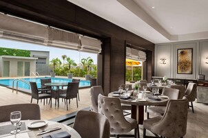 Un moderno comedor con vista a la piscina, mesas dispuestas con platos y vasos, y un amplio ventanal con persianas.
