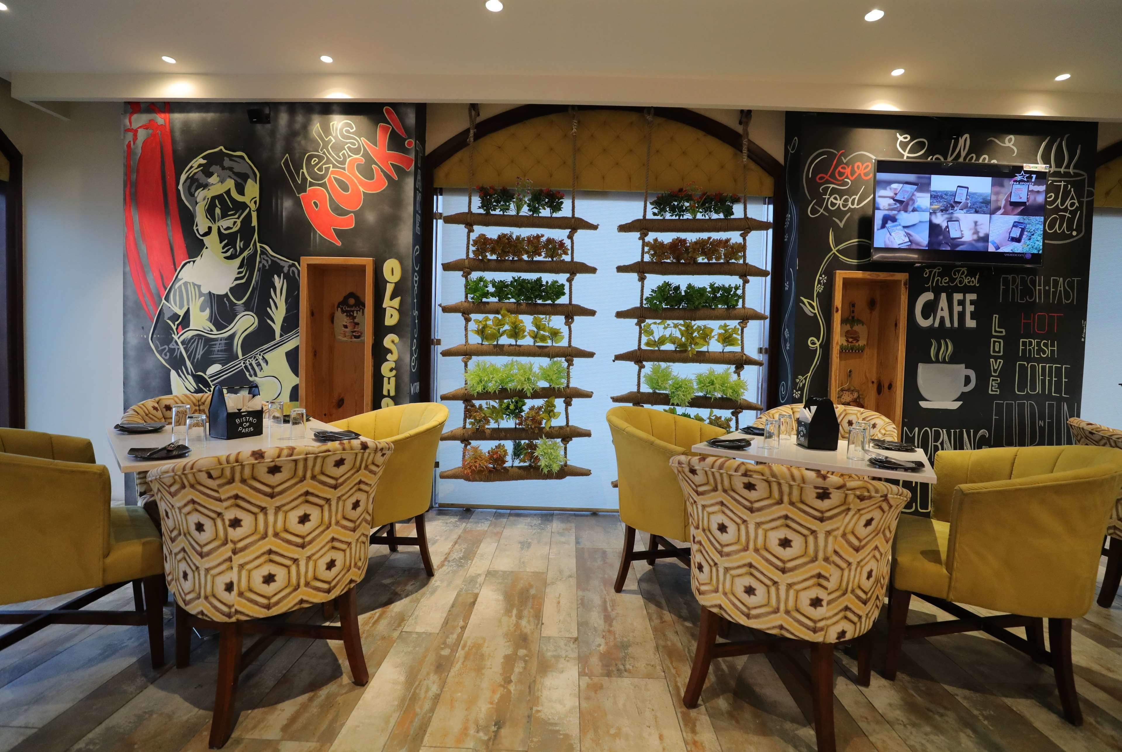 A cafe with yellow chairs, a wall mural, a TV, and a plant shelf.