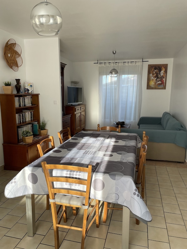 A dining area with a table set for six, surrounded by wooden chairs. A bookshelf with various items is to the left, and a sofa is in the background.