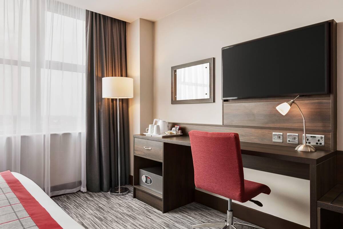 Habitación de hotel con cama, escritorio con silla, televisor, lámpara y ventana con cortinas.