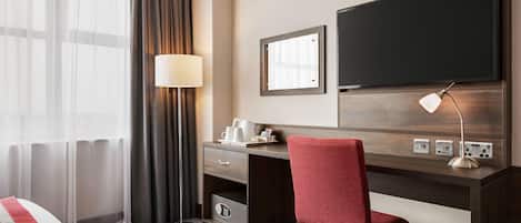 A hotel room with a bed, a desk with a chair, a TV, a lamp, and a window with curtains.