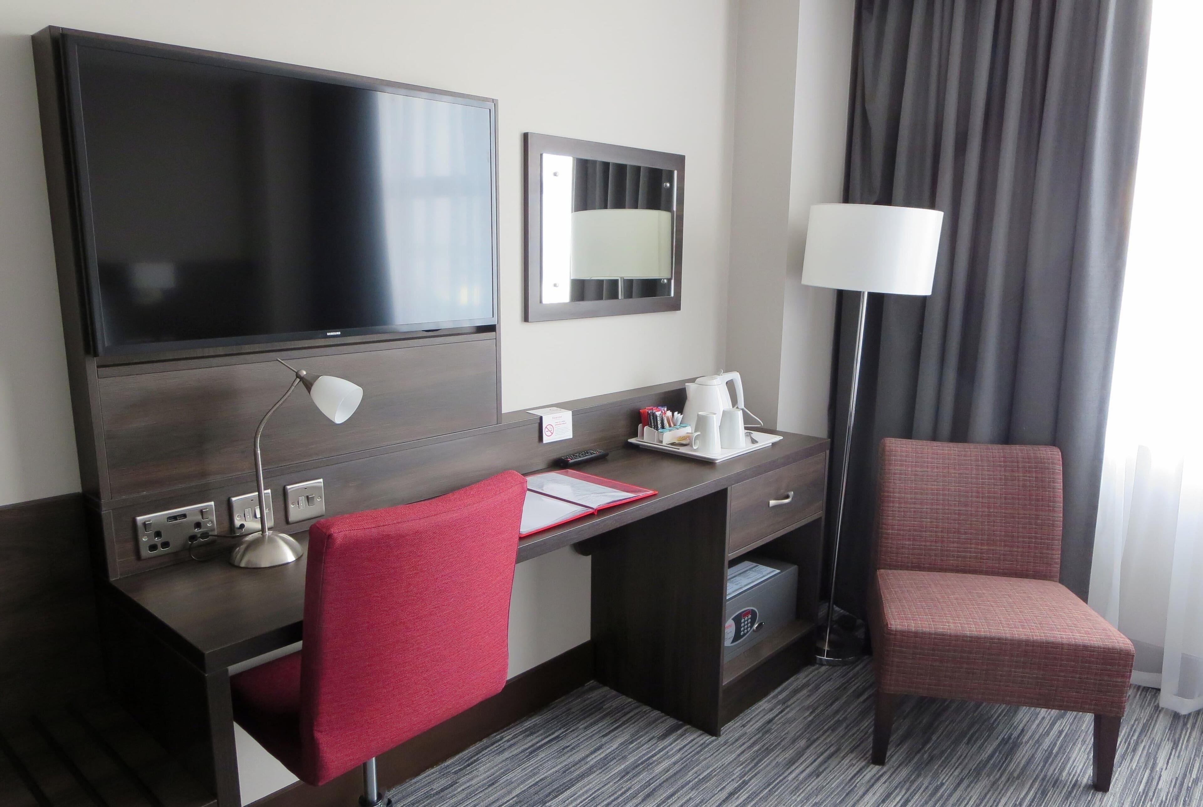 A hotel room with a large flat-screen TV, a desk with a lamp, a red chair, a bedside table with a lamp, and a window with curtains.