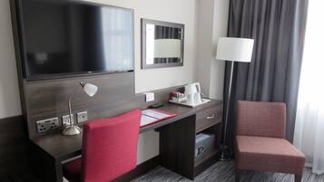 A hotel room with a large flat-screen TV, a desk with a lamp, a red chair, a bedside table with a lamp, and a window with curtains.