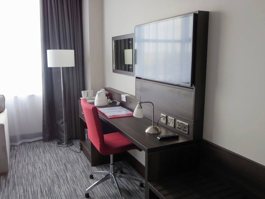 Ein Hotelzimmer mit Schreibtisch, einem roten Sessel, einer Lampe, einem Flachbildfernseher und einem Bett.