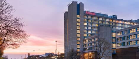 A modern hotel with a prominent 'Ramada' sign, featuring a multi-story structure with numerous windows and a well-lit facade.