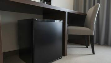 Un petit réfrigérateur noir, un bureau en bois foncé et une chaise rembourrée de couleur grise dans une chambre d’hôtel.
