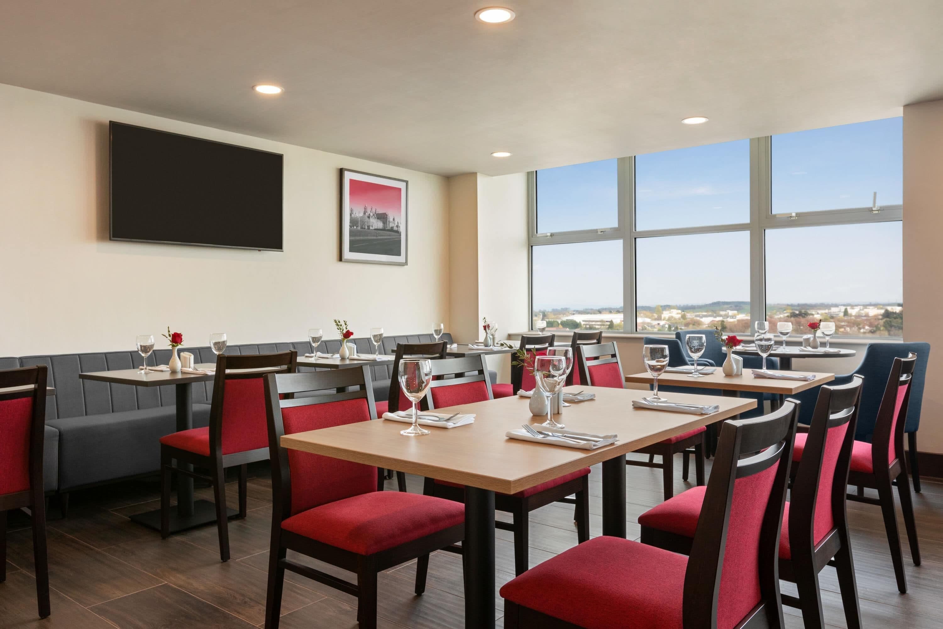A restaurant with multiple tables and chairs, a flat-screen TV mounted on the wall, and large windows offering a view of the city.