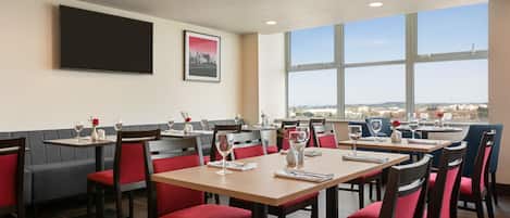 A restaurant with multiple tables and chairs, a flat-screen TV mounted on the wall, and large windows offering a view of the city.