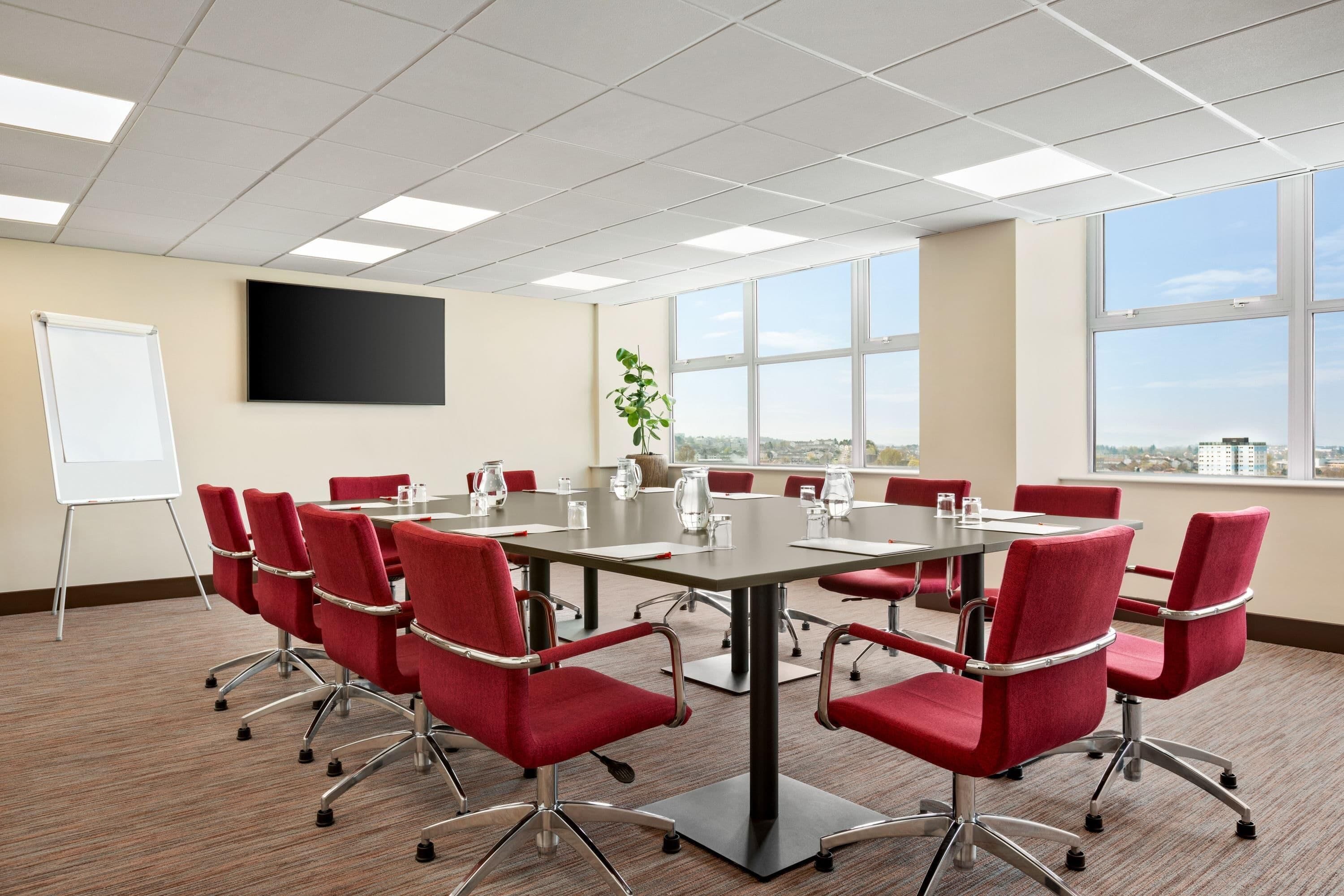 A conference room with a large table, red chairs, a whiteboard, and large windows with a city view.