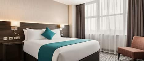 Habitación de hotel con una cama grande, una mesita de noche, una silla y una ventana con cortinas translúcidas.