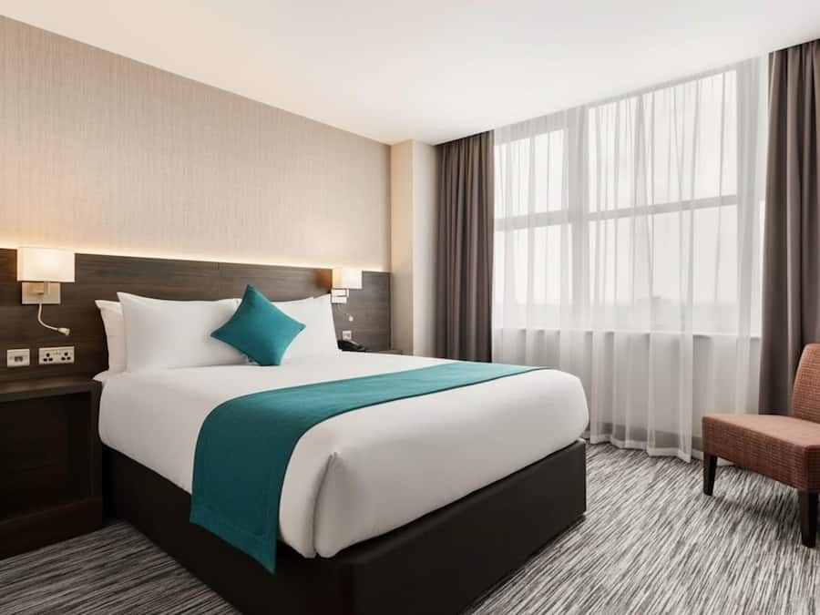 Habitación de hotel con una cama grande, una mesita de noche, una silla y una ventana con cortinas translúcidas.