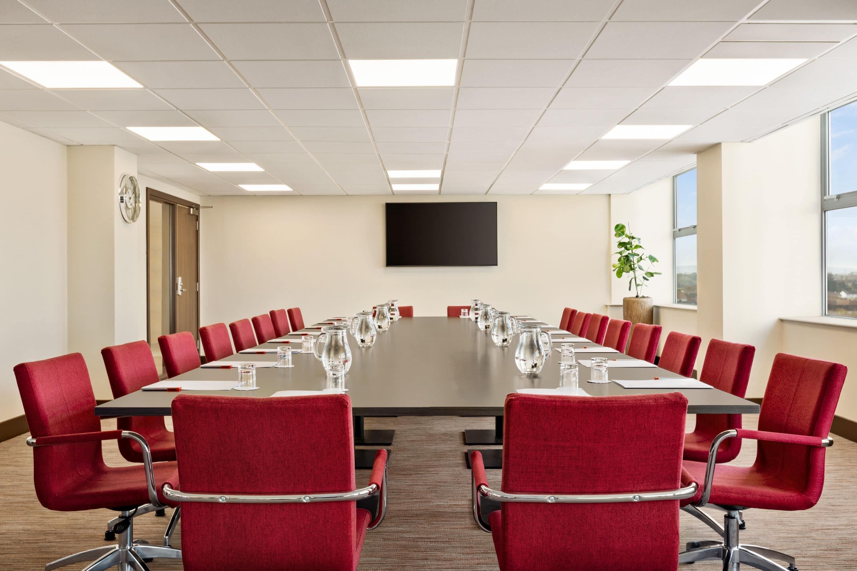 A conference room with a long table, red chairs, a clock, and a TV screen.