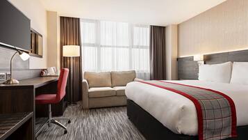 Quarto de hotel com cama, escrivaninha, cadeira, sofá e uma janela grande com cortinas.