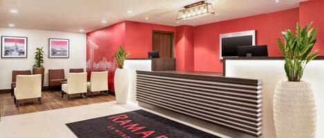 A reception area with a red wall, a reception desk, and a black rug with "RAMADA" on it. There are chairs and potted plants.