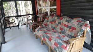 A wooden cabin with a wooden sofa covered in a patchwork quilt, a wooden table with chairs, and a large window with red curtains. - Rustic log cottage, River-view, Pet-friendly, Downeast Maine (Edmunds Township)
