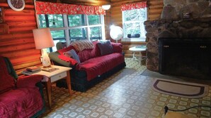 A cozy cabin interior with a wooden log wall, a red sofa, a small table with a lamp, and a stone fireplace. - Rustic log cottage, River-view, Pet-friendly, Downeast Maine (Edmunds Township)