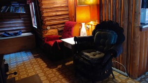 A cozy log cabin room with a wooden wall, a red chair, a black armchair, a table, and a lamp. - Rustic log cottage, River-view, Pet-friendly, Downeast Maine (Edmunds Township)
