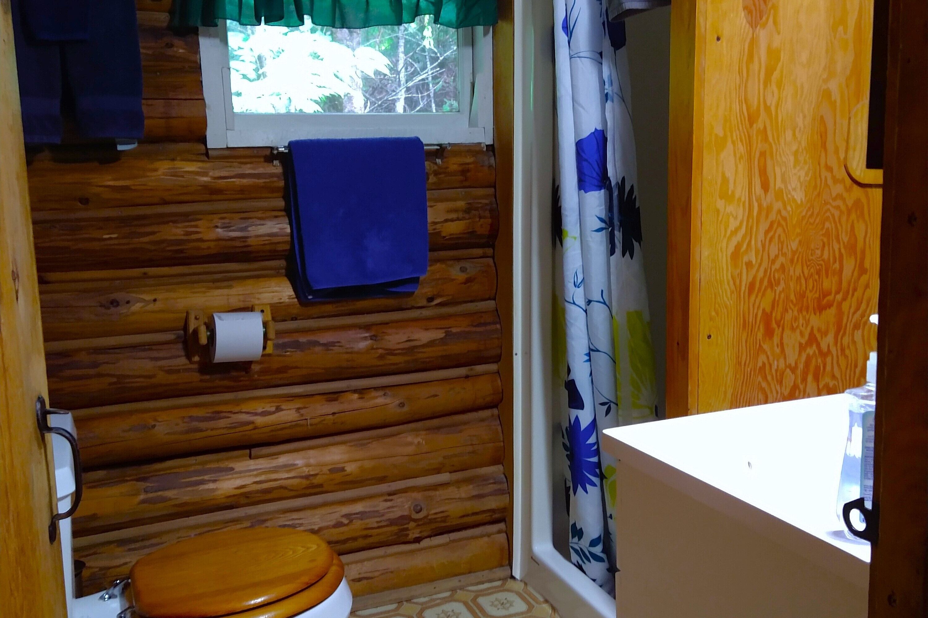 Een houten badkamer in een hutje, met een toilet, een raam met gordijn en een handdoek aan de muur.