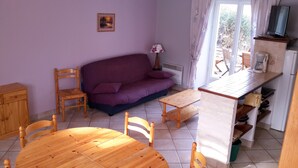 A cozy living area with a purple sofa, wooden chairs, a small wooden table, and a kitchen area with a TV and coffee maker. - Apartment with large garden prox. from the sea and landing beaches (Étréham)