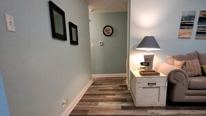 Un couloir au plancher de bois menant à un espace de vie comprenant un canapé et une lampe.