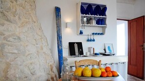 A kitchen with a stone wall, a wooden table with a fruit plate, and a shelf with kitchenware.