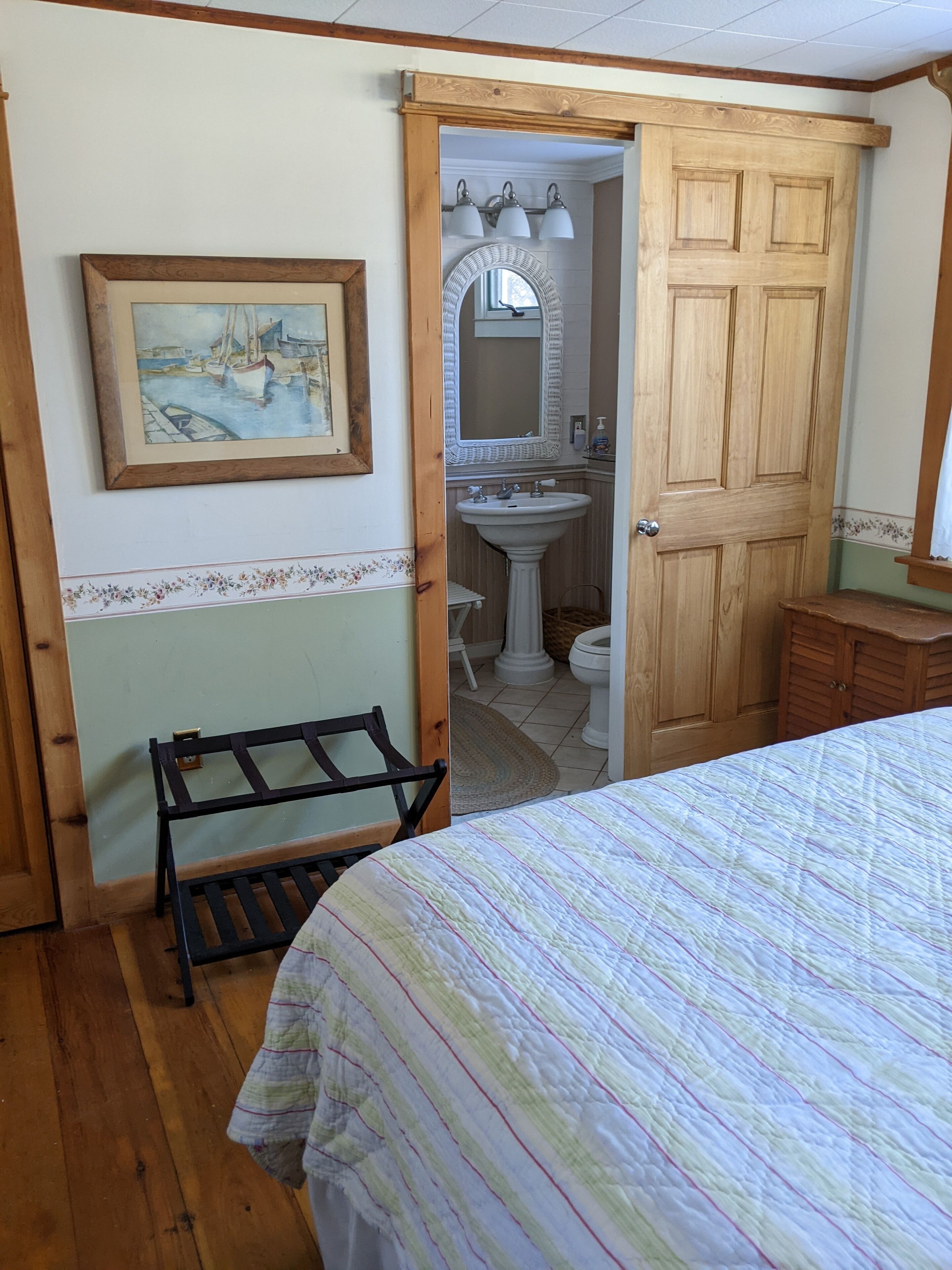 A bedroom with a bed, a wooden bench, a painting, a bathroom with a sink and mirror, and a wooden door.