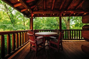 A wooden deck with a round table and chairs, surrounded by a wooden railing, overlooking a forested area.