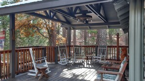A covered outdoor deck with wooden furniture, including rocking chairs and benches, surrounded by trees.