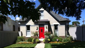Une maison à deux étages avec une porte d’entrée rouge, un petit jardin devant la maison agrémenté de différentes plantes et une antenne parabolique bien visible.