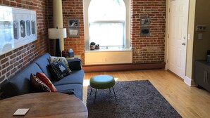 Un salon chaleureux avec un mur en briques, un canapé bleu, une table basse en bois, un pouf vert et une grande fenêtre avec des stores.