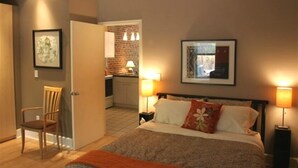 Une chambre à coucher comprenant un lit, des lampes de chevet, une chaise, une porte donnant sur la cuisine et des œuvres d’art encadrées accrochées aux murs.